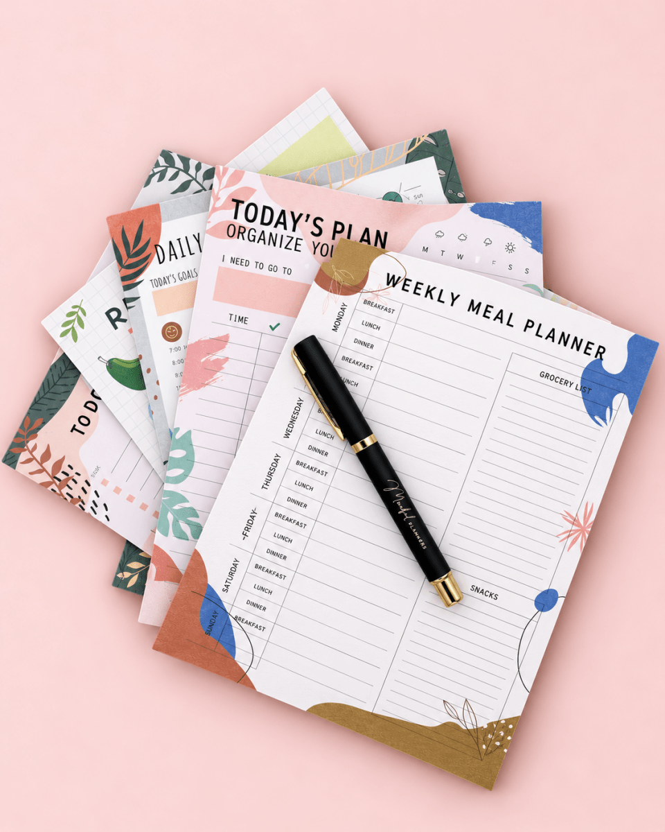 A styled flat-lay of a paper planner set on a soft pink background. The set includes a weekly meal planner pad, a daily planner pad, and other coordinating planner sheets with pastel botanical and geometric designs.