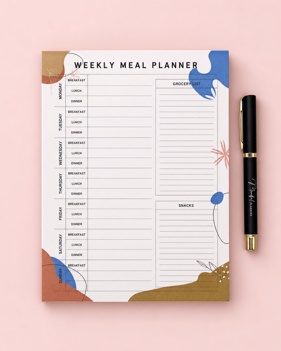 A weekly meal planner pad with a clean white background and colorful abstract accents in blue, brown, and gold, shown next to a black pen with gold trim on a solid pink background.